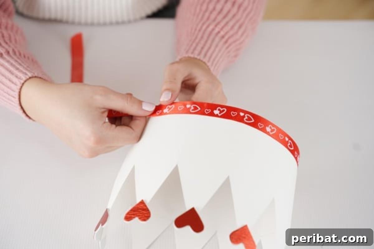 Easy Valentine Paper Crown Craft for Children 12 A hand adheres a festive ribbon along the bottom edge of the paper crown, creating a decorative trim.