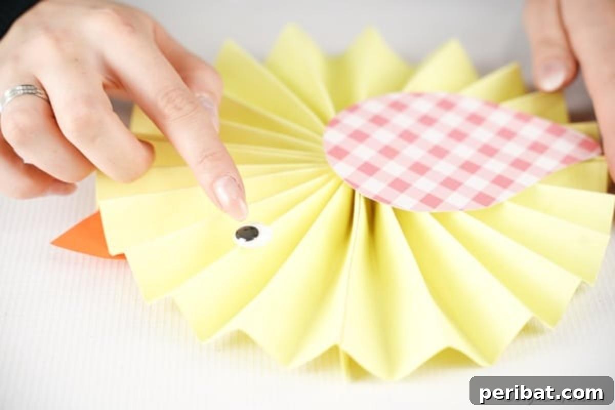 Simple & Sweet: Crafting Paper Birds with Kids 14 A close-up of a hand attaching a googly eye and an orange beak to the yellow paper fan bird, carefully bringing its face to life with these final details.