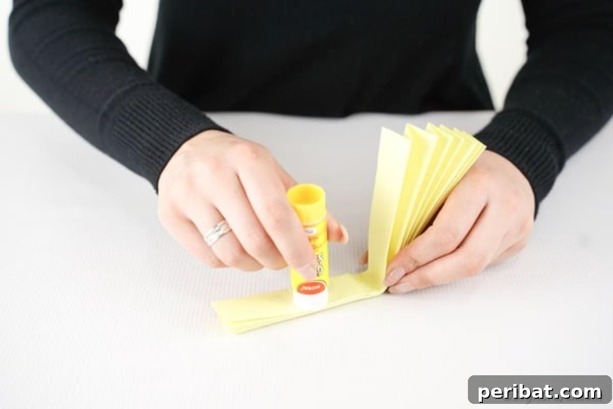 Simple & Sweet: Crafting Paper Birds with Kids 9 A close-up shot of hands applying glue stick to the inside edge of the folded paper fan, preparing to securely attach the two sides to form the circular bird body.