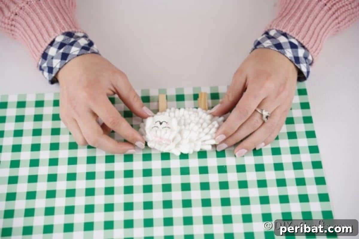 Attaching the paper lamb body to two clothespins, which will serve as its legs, with craft glue, ensuring stability.