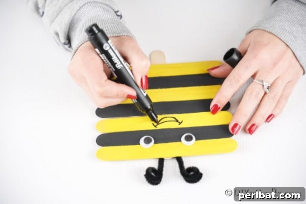 Googly eyes being added and a smile being drawn on the craft stick bee, giving it a friendly face.