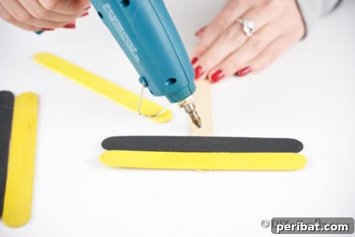 The process of gluing the first yellow craft stick horizontally onto an unpainted stick, demonstrating the initial assembly.
