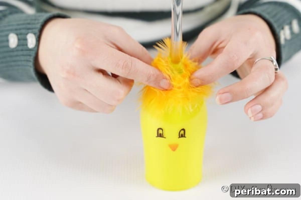 Adorable DIY Chick Wine Glass Luminary 11 Close-up of hands carefully gluing yellow feathers onto the top of a painted wine glass, completing the chick's fluffy look.