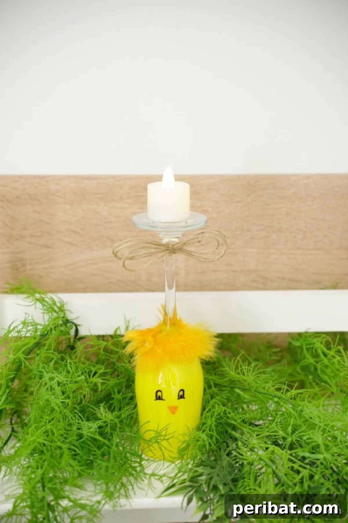 Adorable DIY Chick Wine Glass Luminary 7 A painted wine glass chick candle holder perched on fake grass, framed by a rustic wooden fence, evoking a pastoral Easter scene.