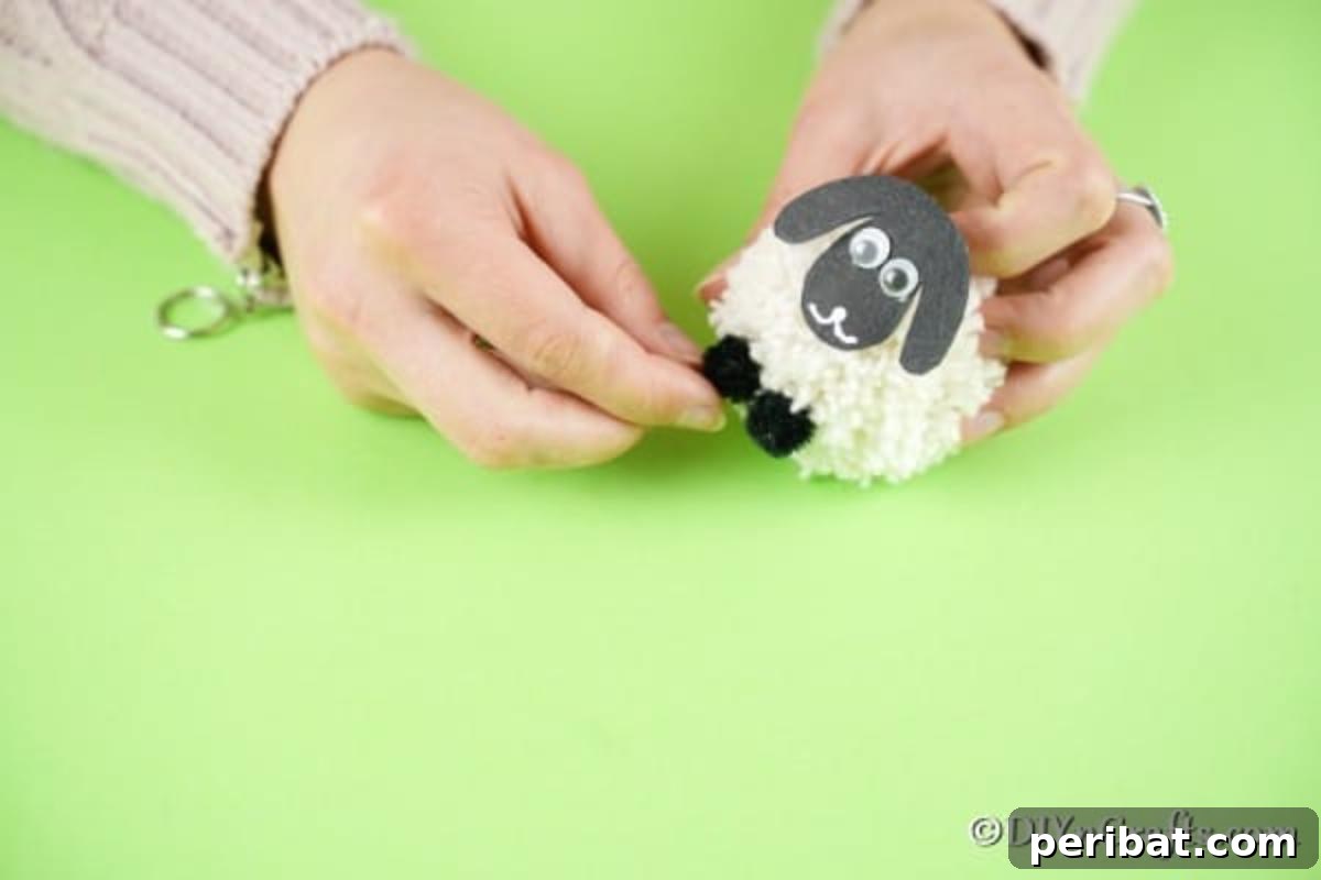 Fluffy Pom-Pom Sheep: A DIY Video Craft Guide 14 Hands carefully gluing a black paper sheep face onto the fluffy yarn pom pom body, and then attaching two small black pom poms underneath to serve as the sheep's feet, completing the assembly.