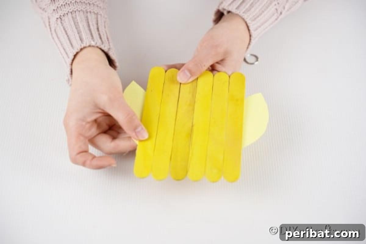 Colorful Craft Stick Chicken Painting 12 Hands carefully gluing a yellow paper wing onto the side of the craft stick chicken body.