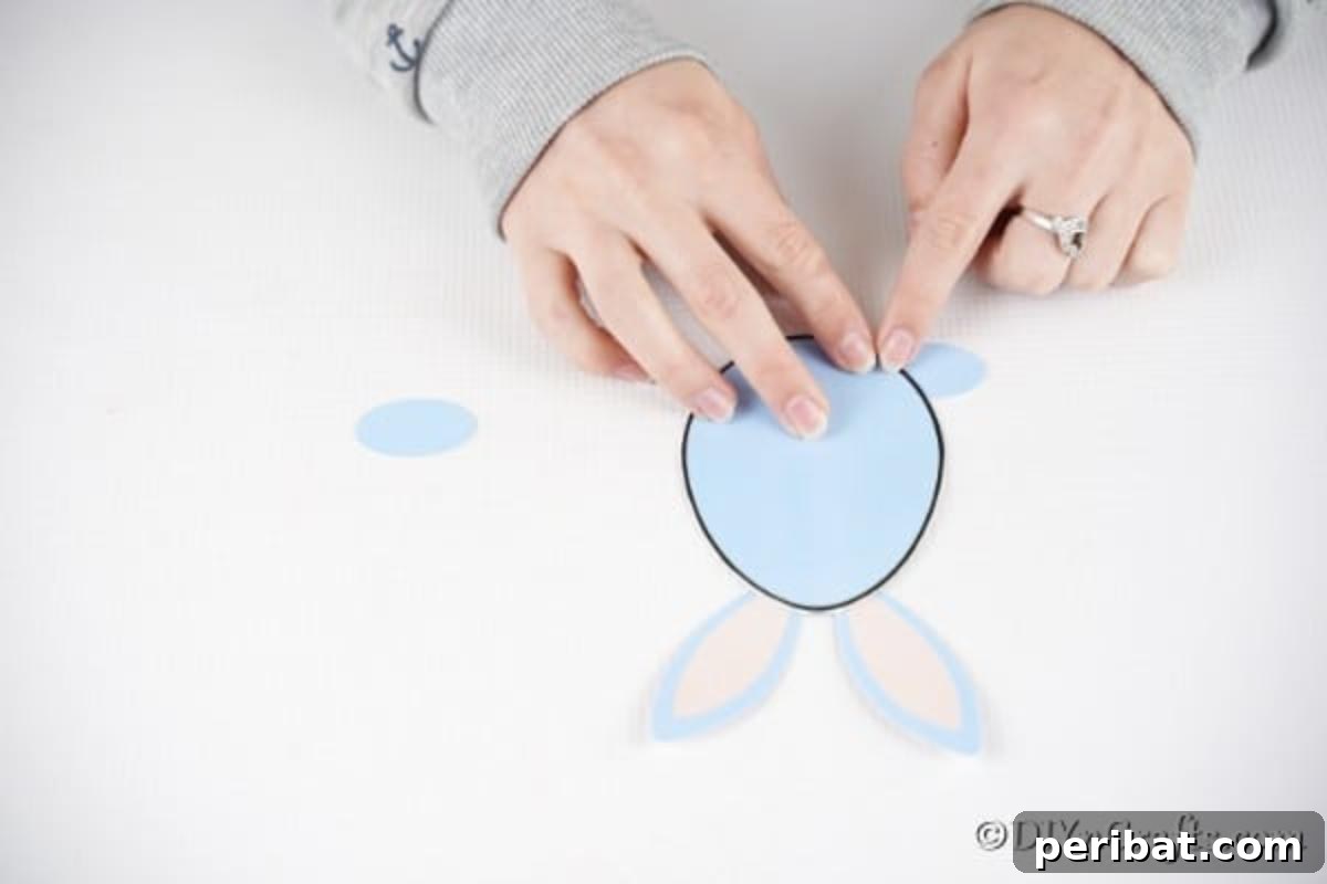 Hands gluing the foot shapes to the bottom edge of the egg-shaped card, completing the bunny's lower body.