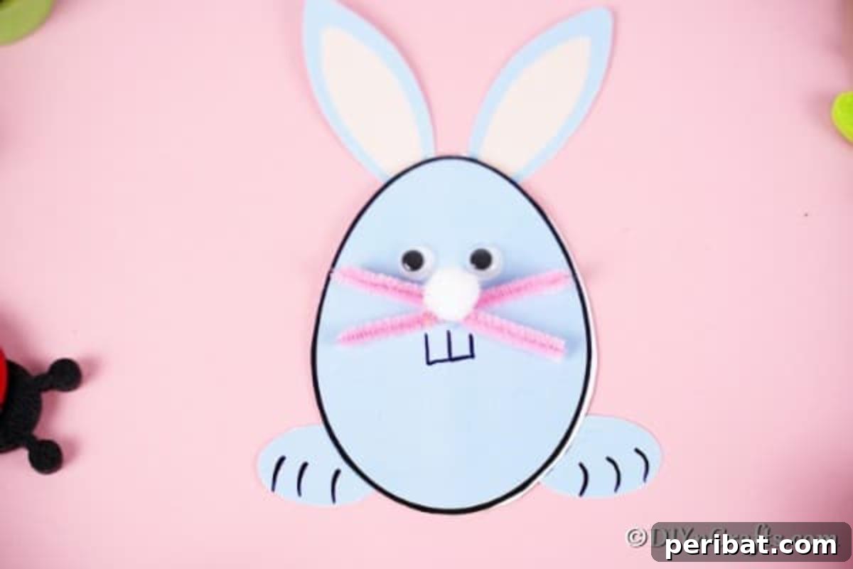 A finished egg shaped Easter bunny card with googly eyes and pipe cleaner whiskers, placed on a soft pink surface.