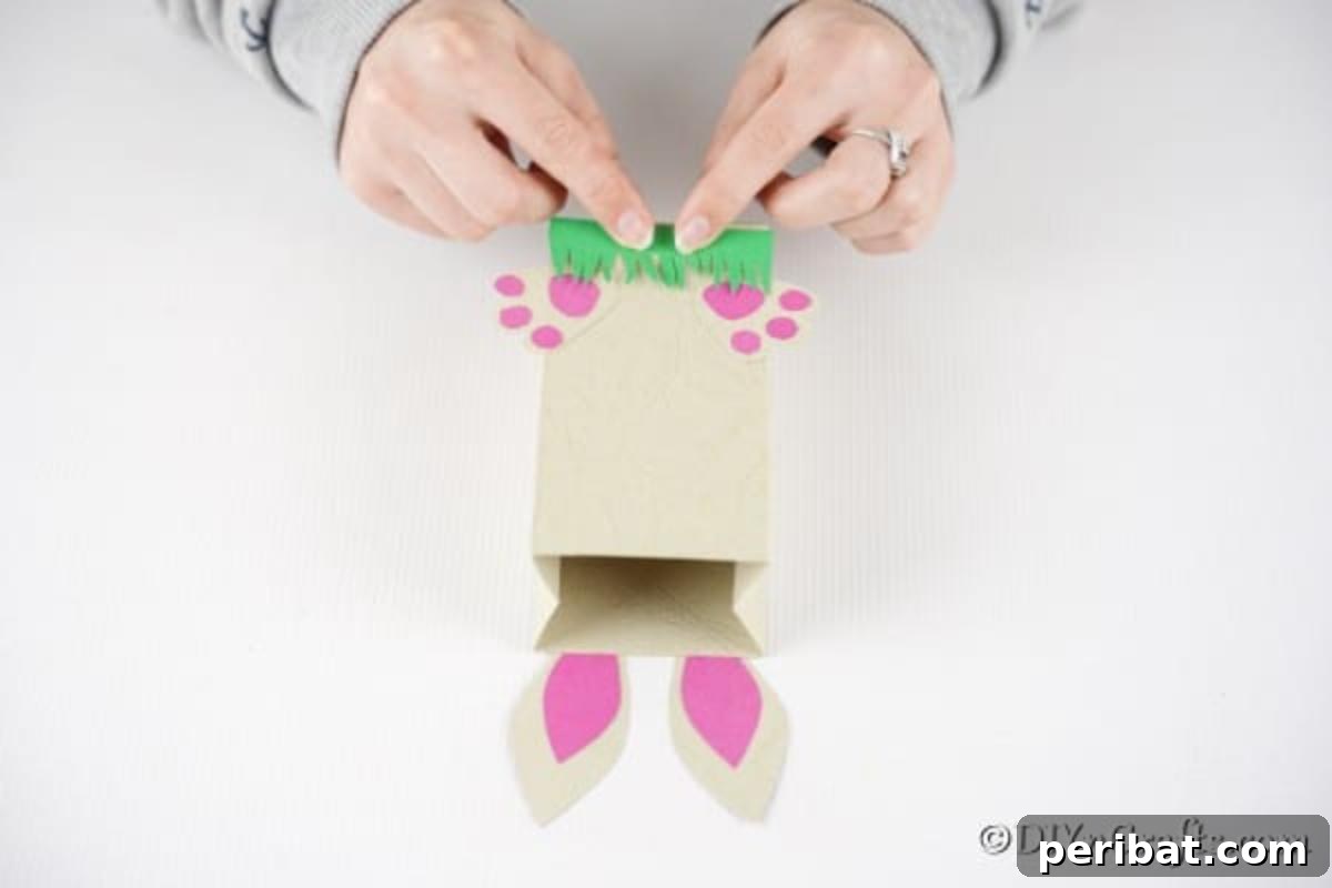 DIY Easter Treat Pouch Video Guide 19 A hand gluing the green grass fringe onto the bottom of the paper bag, beneath the bunny feet
