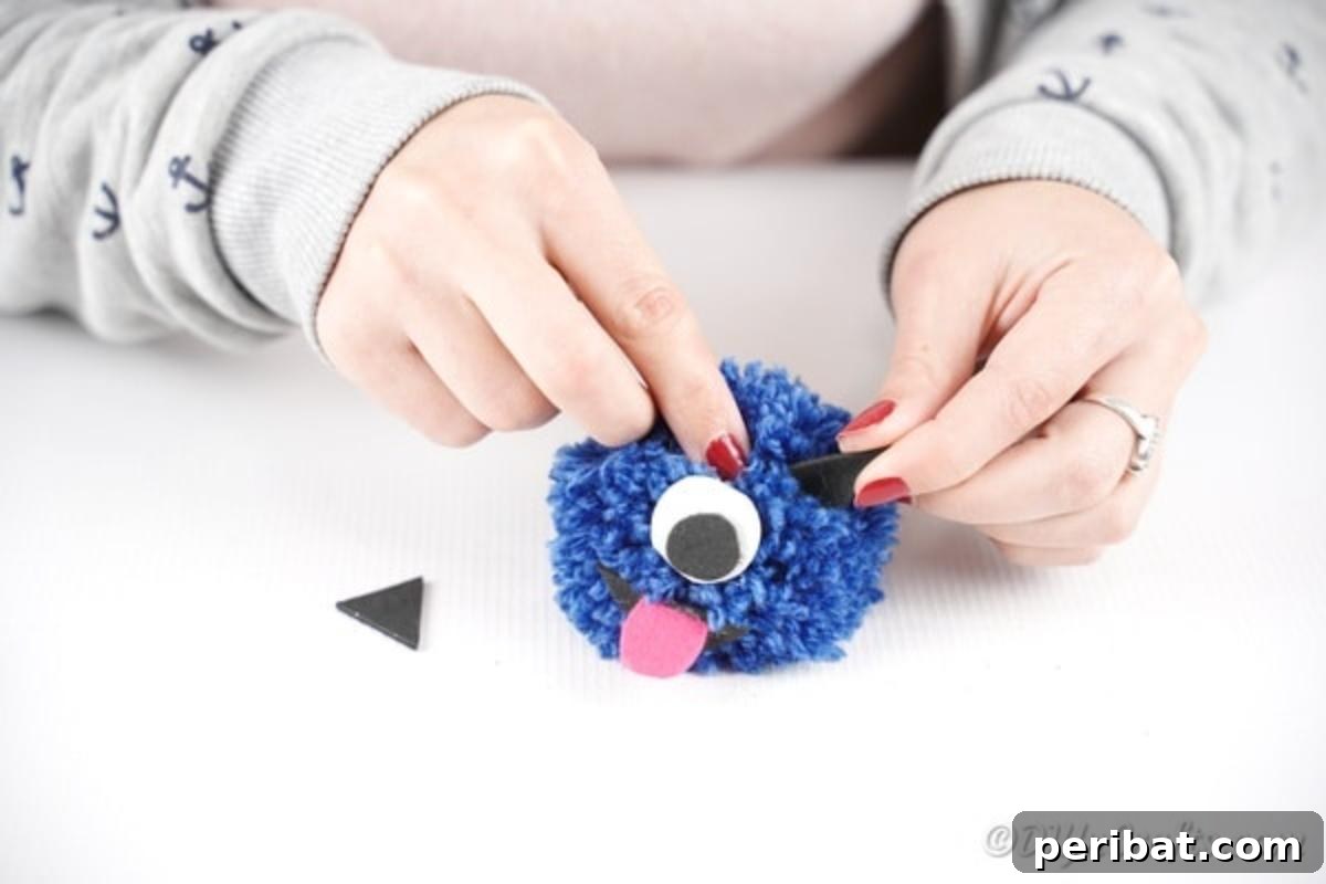Black paper ears being glued onto the sides of the blue pom pom monster