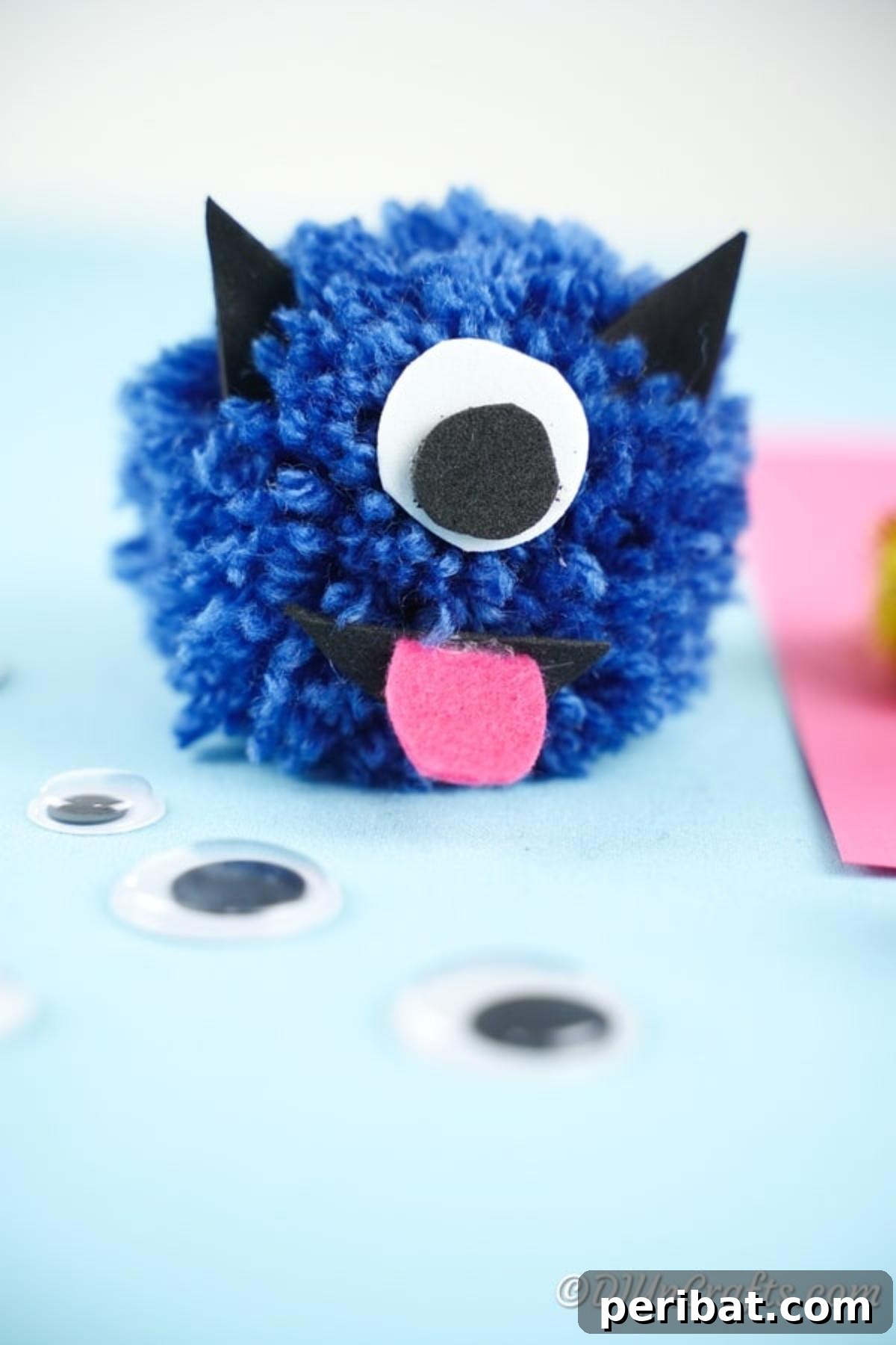 Bright blue yarn pom pom monster with a pink tongue, one large eye, and black ears on a blue background
