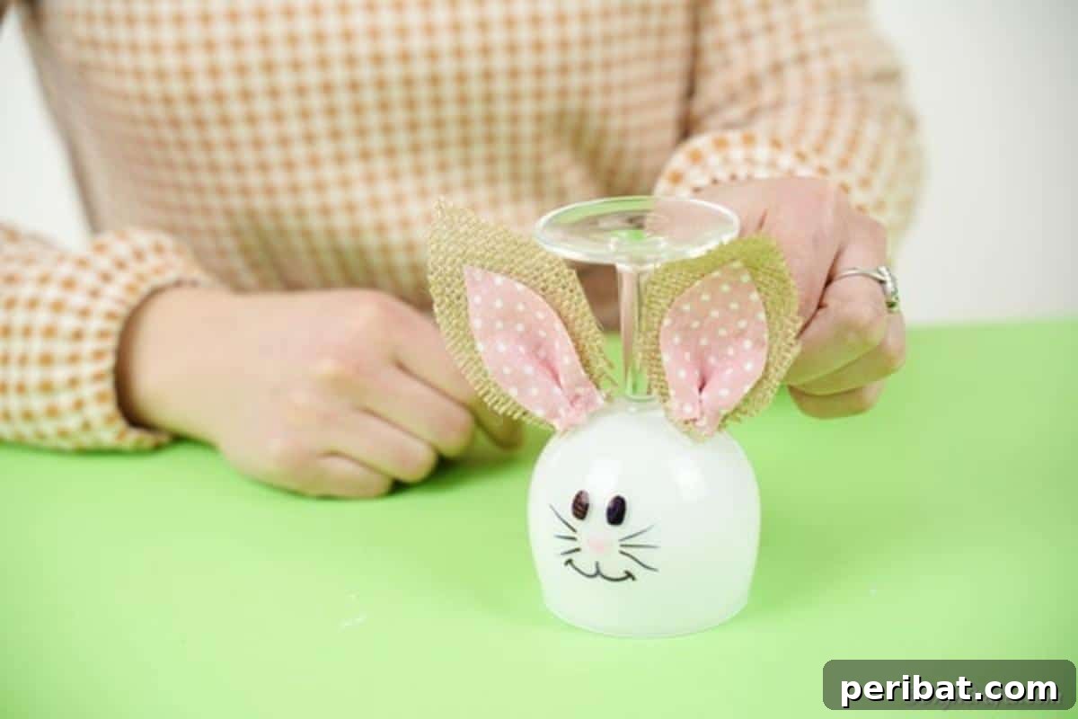 Hoppy Wine Glass Bunny Decor Tutorial 15 A hand using a hot glue gun to attach the burlap and fabric ears to the top of the wine glass.