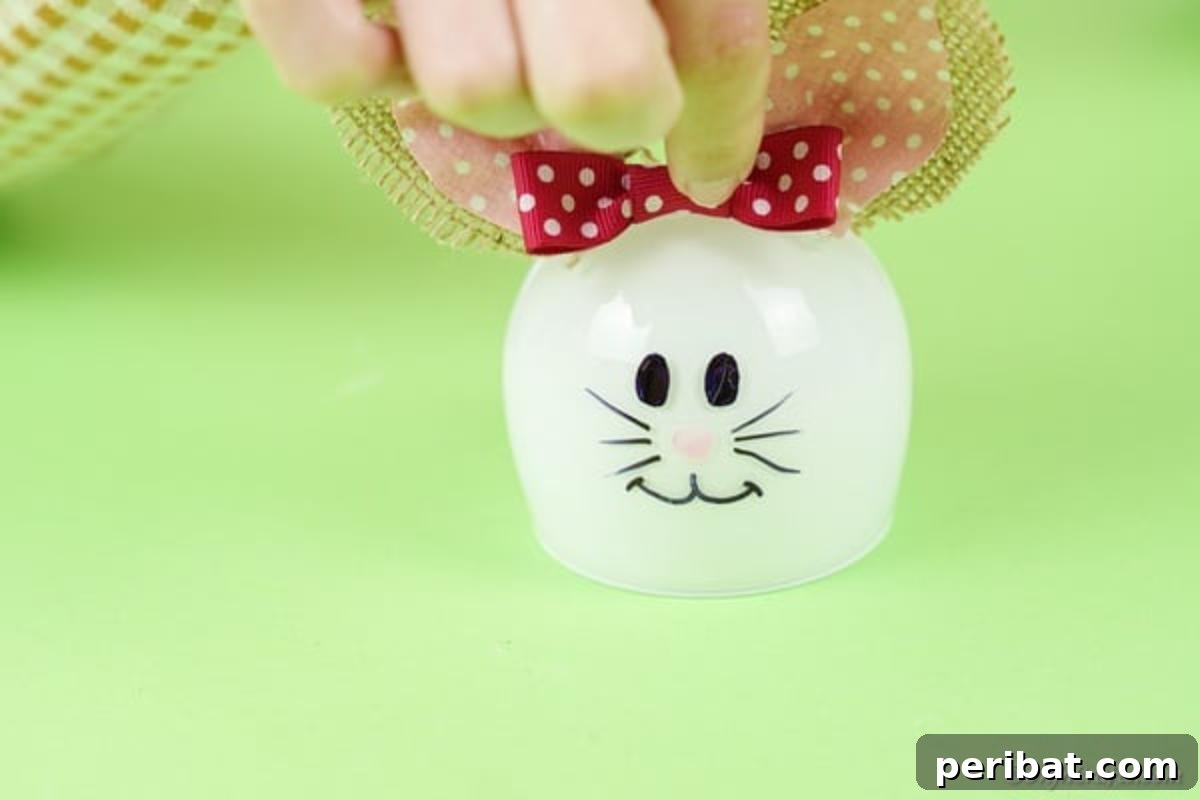 Hoppy Wine Glass Bunny Decor Tutorial 16 A hand adding a small ribbon bow to the top of the wine glass bunny, between its ears.