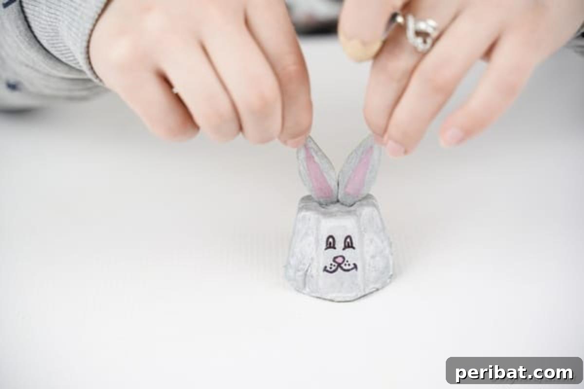 Adorable Upcycled Egg Carton Easter Bunny Craft 13 The final assembly step, where the painted ears are being carefully glued onto the top of the egg carton cup, completing the bunny's silhouette.