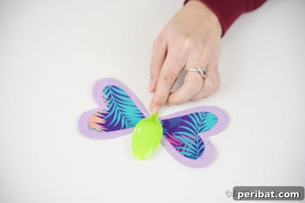 A plastic spoon being carefully glued onto the center of the paper wings, forming the body of the upcycled butterfly craft.