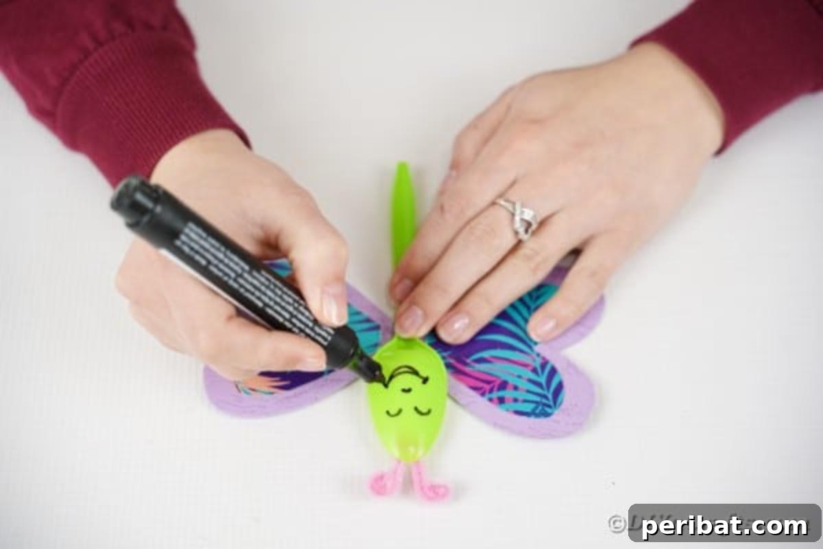 A paint pen being used to draw a cheerful face, complete with eyes and a smile, onto the spoon body of the butterfly.