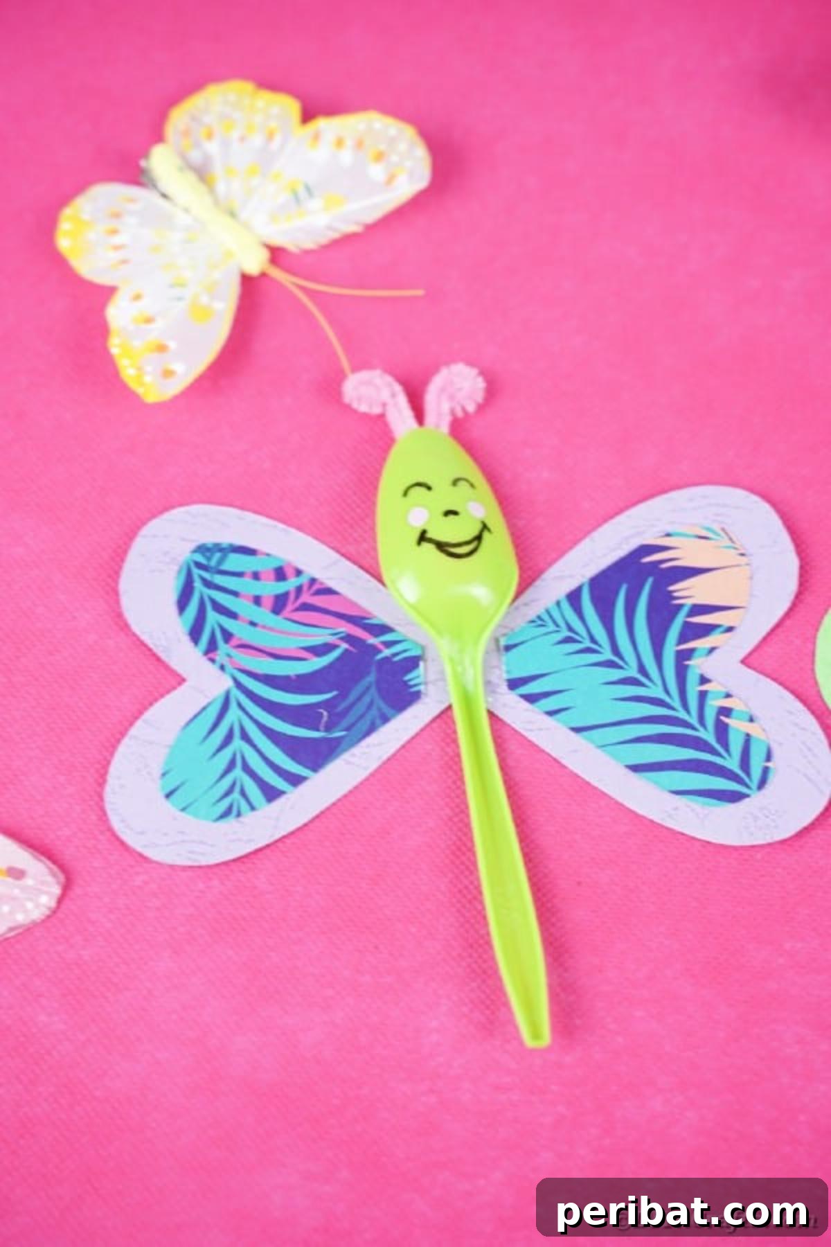 A single green upcycled spoon butterfly with vibrant paper wings resting elegantly on a soft pink paper background, highlighting its delicate details.