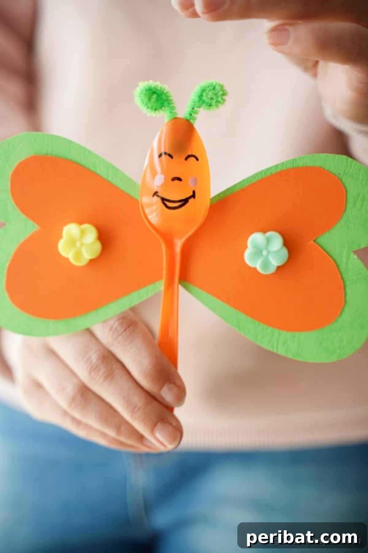 A woman gracefully holds an orange and green upcycled spoon butterfly, highlighting its cheerful design and handcrafted quality.