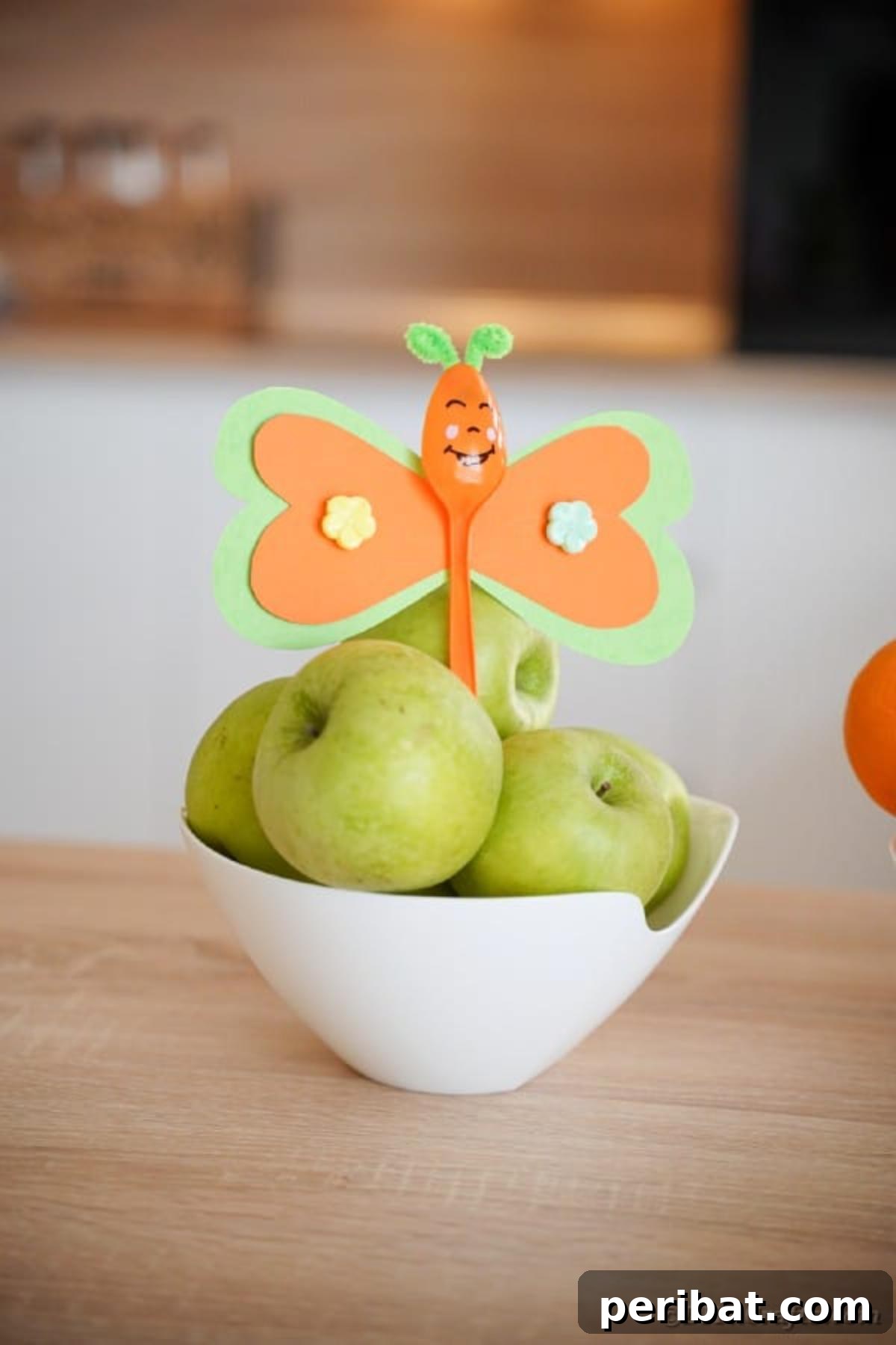 An orange upcycled spoon butterfly perched atop a pile of fresh apples, creating a vibrant and whimsical centerpiece.