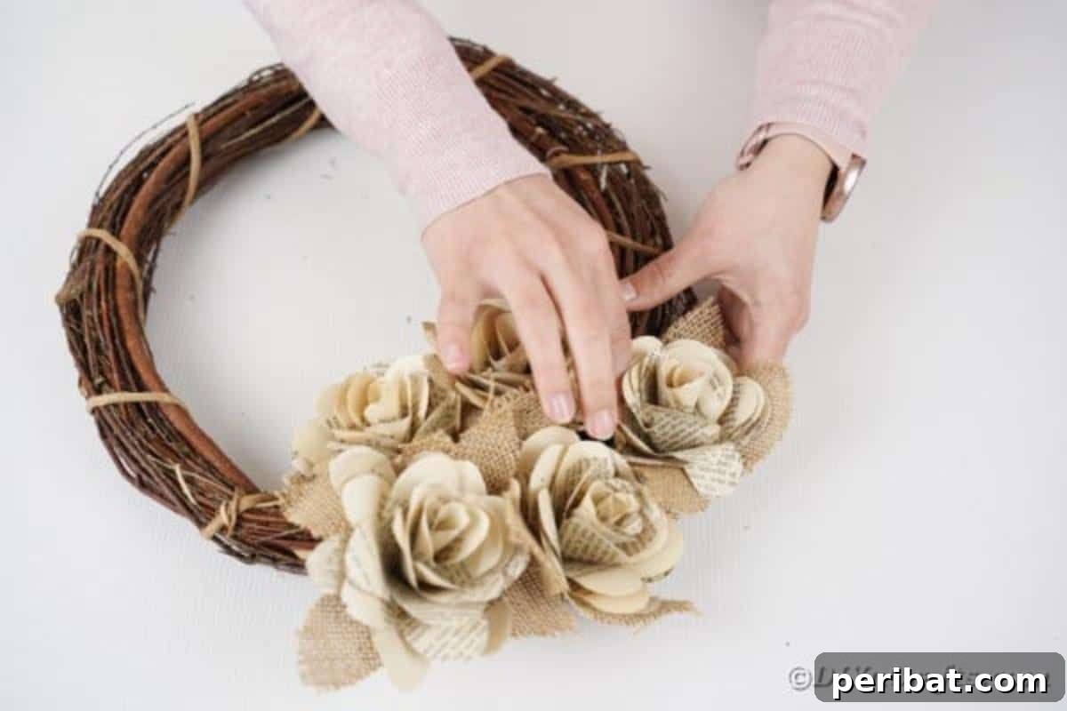 A hand arranging paper roses and burlap leaves on a grapevine wreath, considering placement before final gluing.