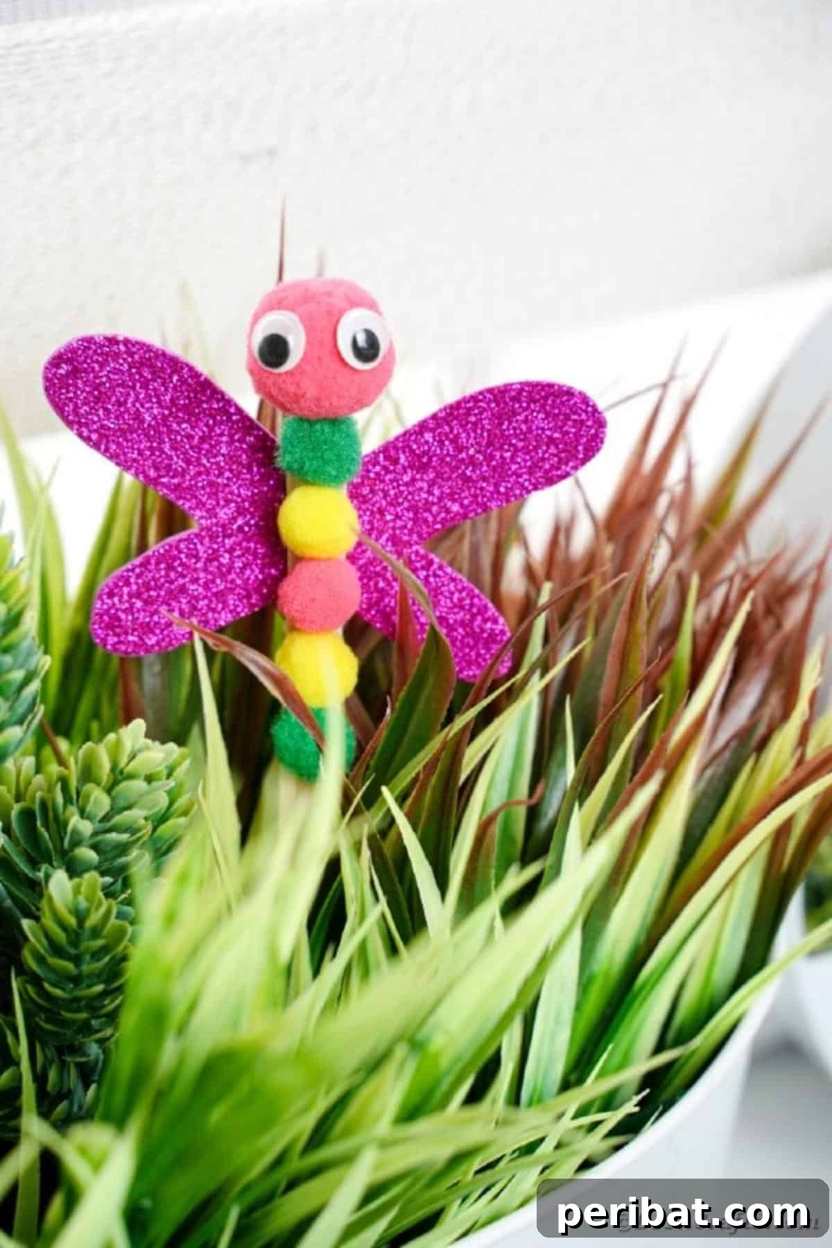 Create Vibrant Craft Stick Dragonflies for Kids 16 Close-up of a DIY craft stick dragonfly craft with colorful pom-pom body and glitter wings, sticking out of a green potted plant.