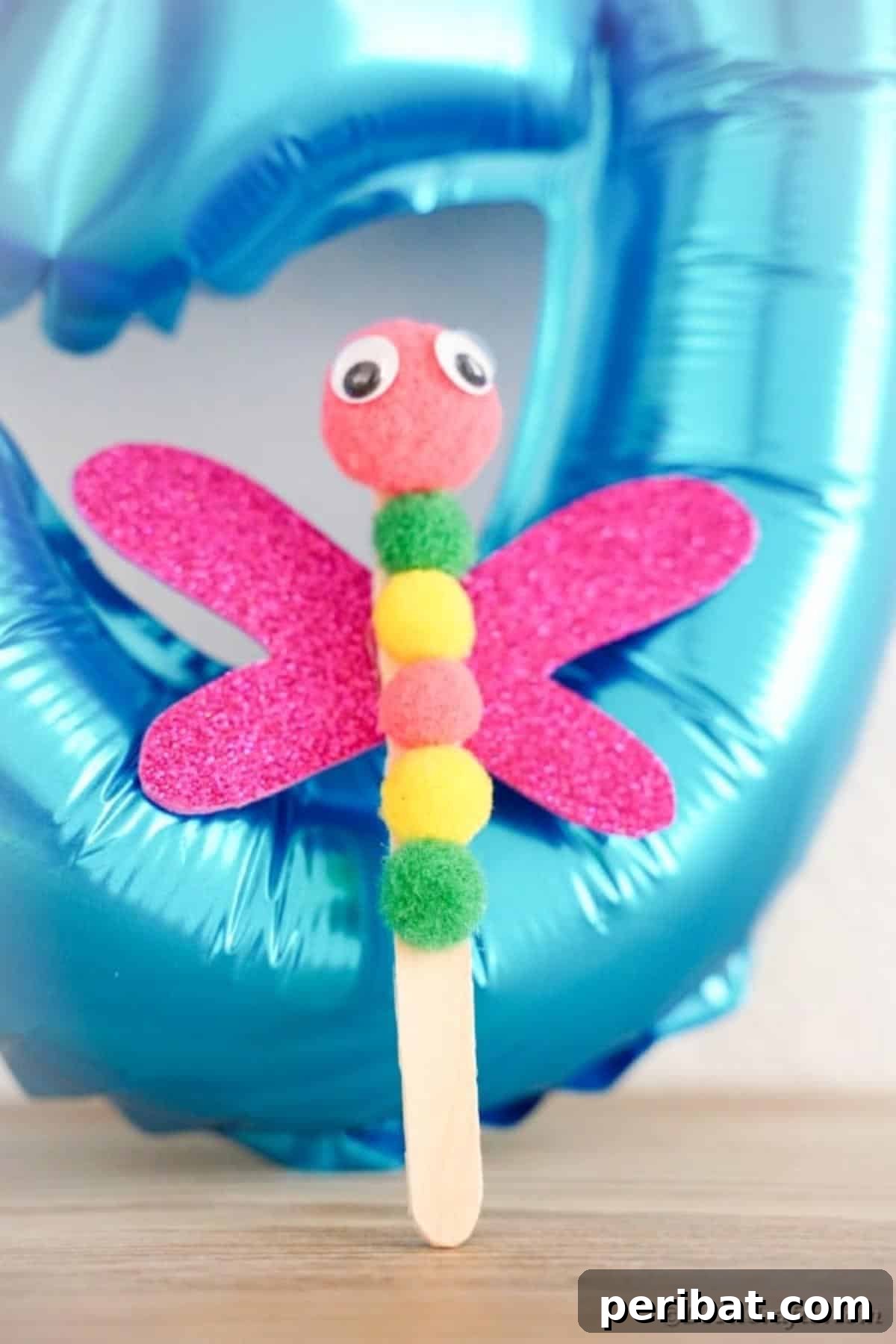 Create Vibrant Craft Stick Dragonflies for Kids 18 A charming craft stick dragonfly held up against a blurred blue background, showcasing its sparkling wings and bright pom-pom body.