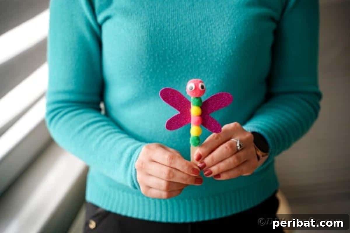 Create Vibrant Craft Stick Dragonflies for Kids 14 A woman in a teal shirt gently holding a colorful craft stick dragonfly, showcasing the completed project with a soft focus background.