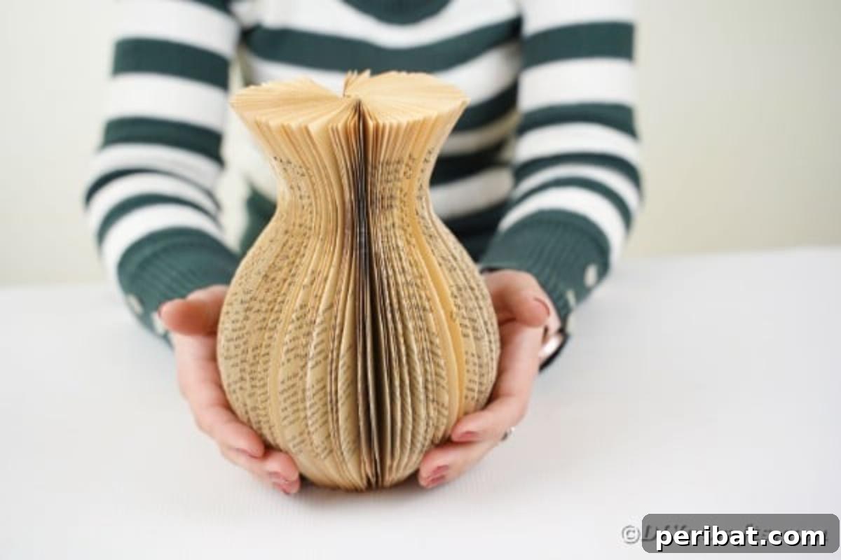 A person holding the fanned-out pages of a cut book, gently pulling the ends together to form the cylindrical shape of a paper vase.