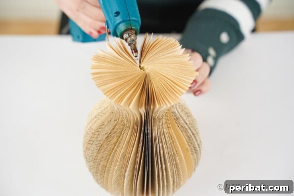 A close-up shot of a person applying hot glue to the final pages of a paper vase made from an old book, securing the circular shape.