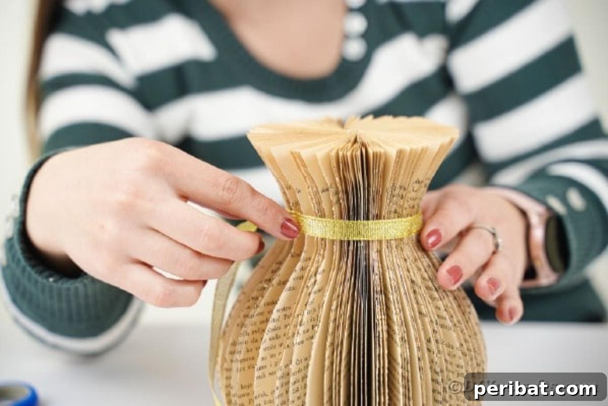 A person carefully gluing a strip of gold ribbon around the neck of a finished paper vase, adding a decorative accent.