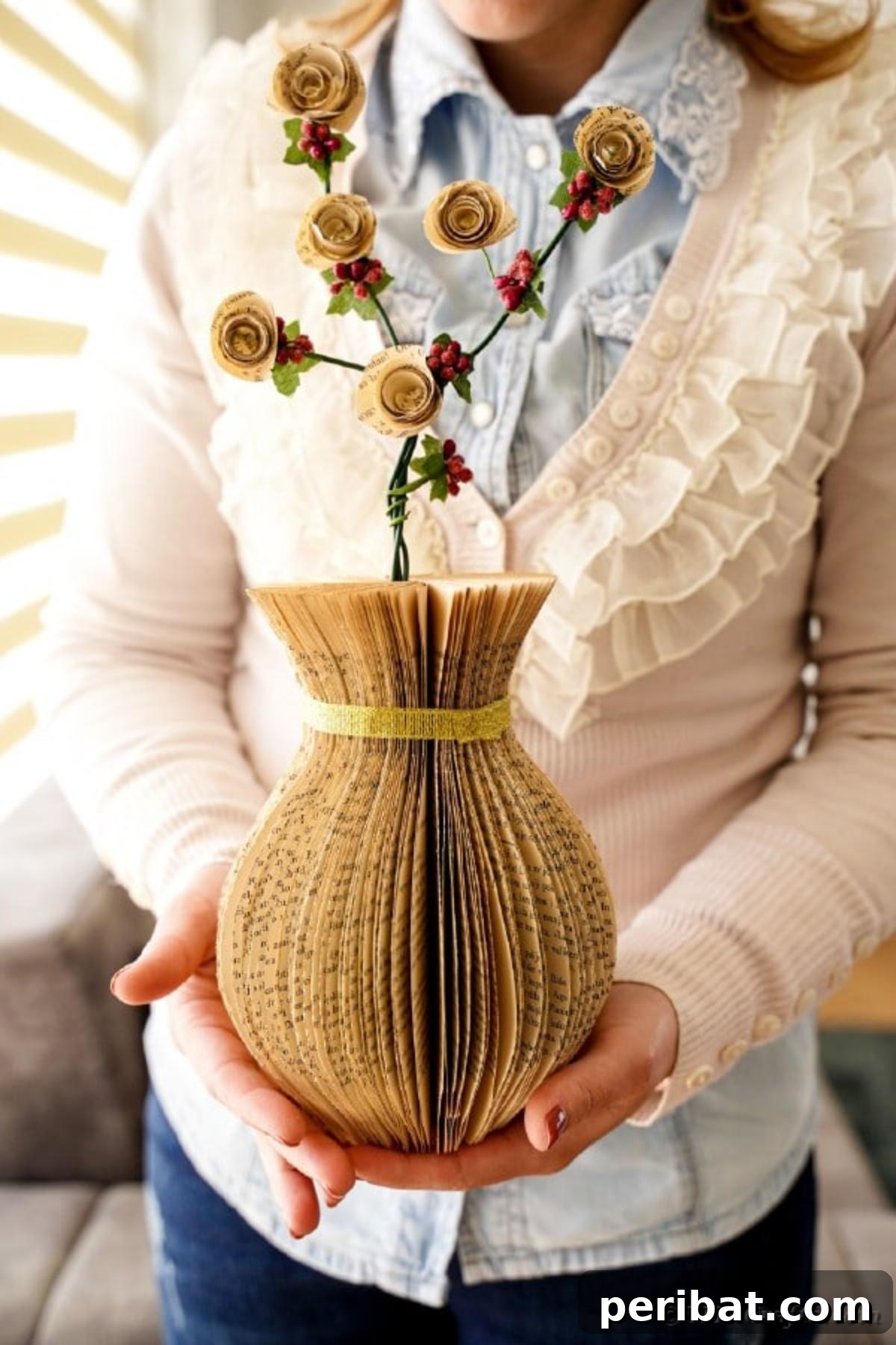 A finished paper vase made from old book pages, held by a woman, adorned with a gold ribbon and filled with artificial flowers, ready for display.