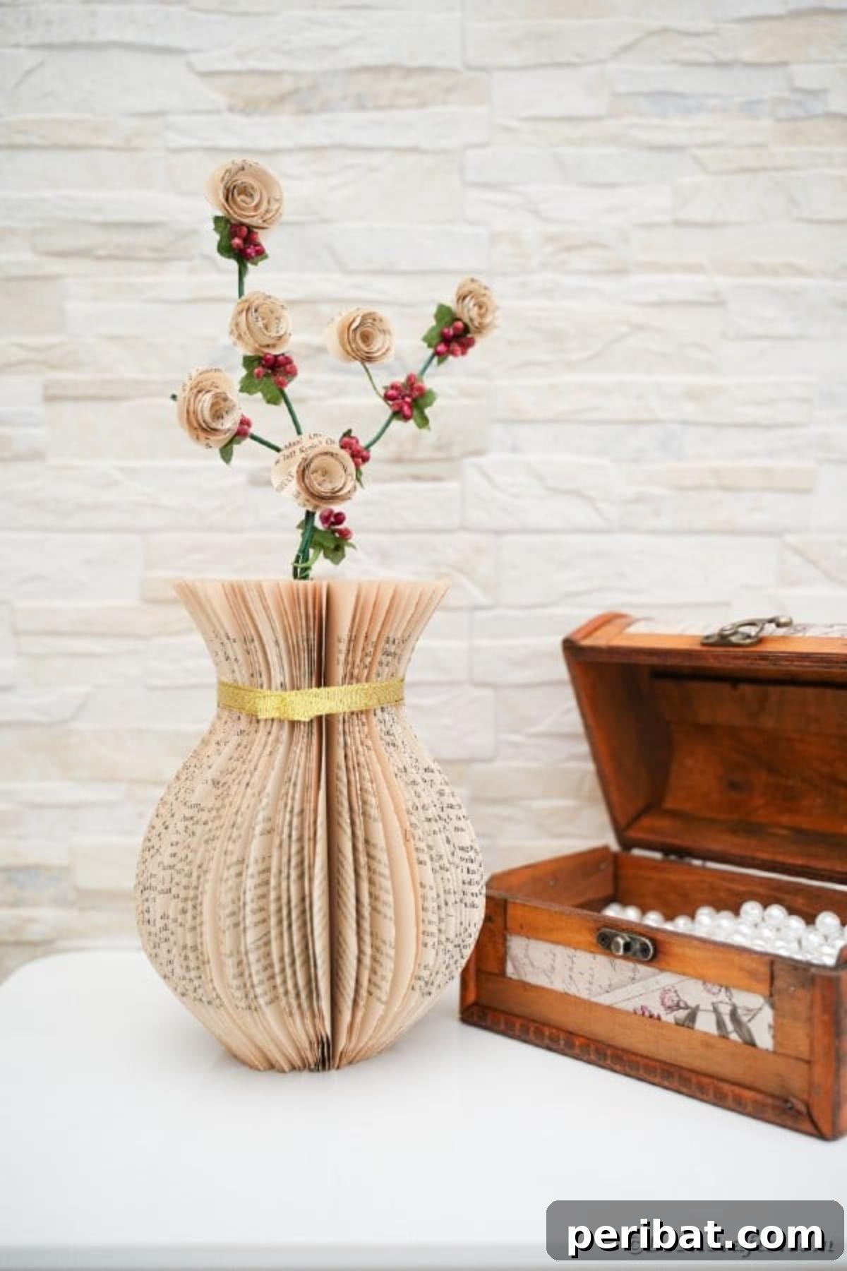 A charming paper vase crafted from old book pages, accented with a gold ribbon, displayed on a wooden table beside an open rustic chest, creating a harmonious blend of textures and styles.