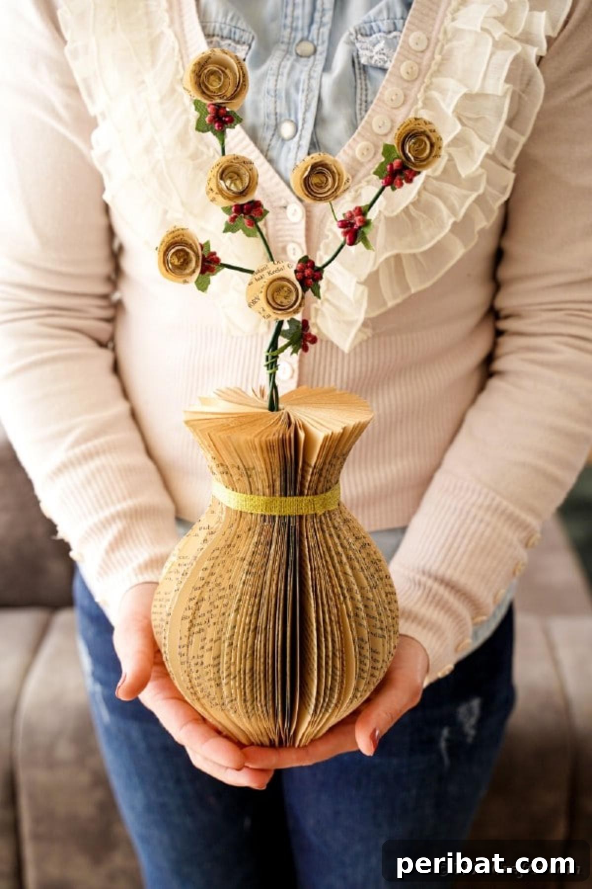 A woman gracefully holding a handcrafted paper vase made from old book pages, featuring a delicate gold ribbon and filled with elegant artificial flowers, exemplifying its charming aesthetic.
