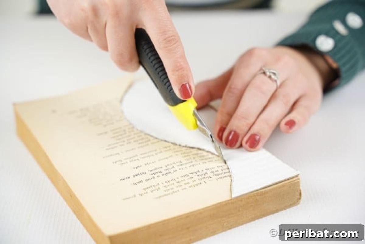 A craft knife carefully cutting through multiple pages of an old book, following a pre-drawn vase outline, illustrating the precision required for the project.