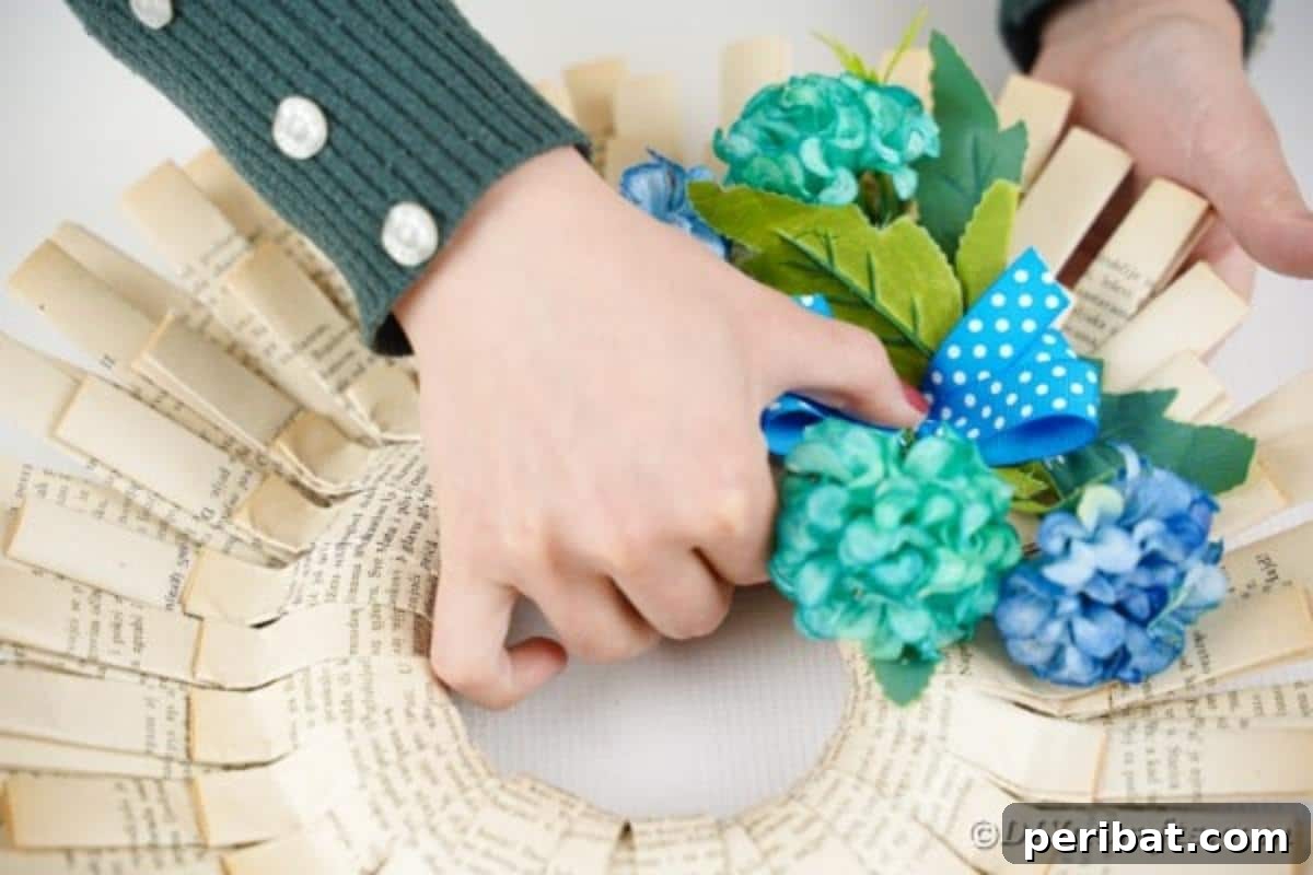 A small blue polka dot bow is being added to the cluster of blue flowers on the paper wreath, completing the decorative arrangement.