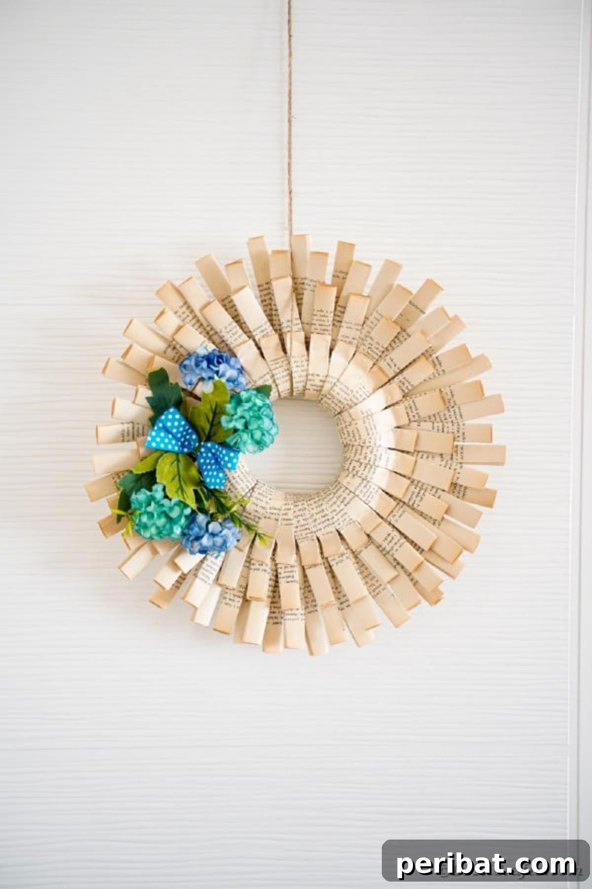 A delicate paper wreath, meticulously crafted from book pages and adorned with soft blue artificial flowers, hangs gracefully on a clean white wall.