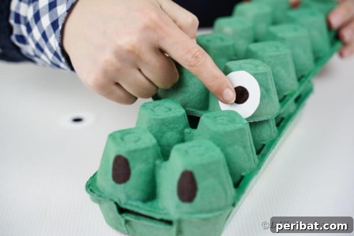 A hand gluing a white paper eye with a black pupil onto the green egg carton dragon's head.