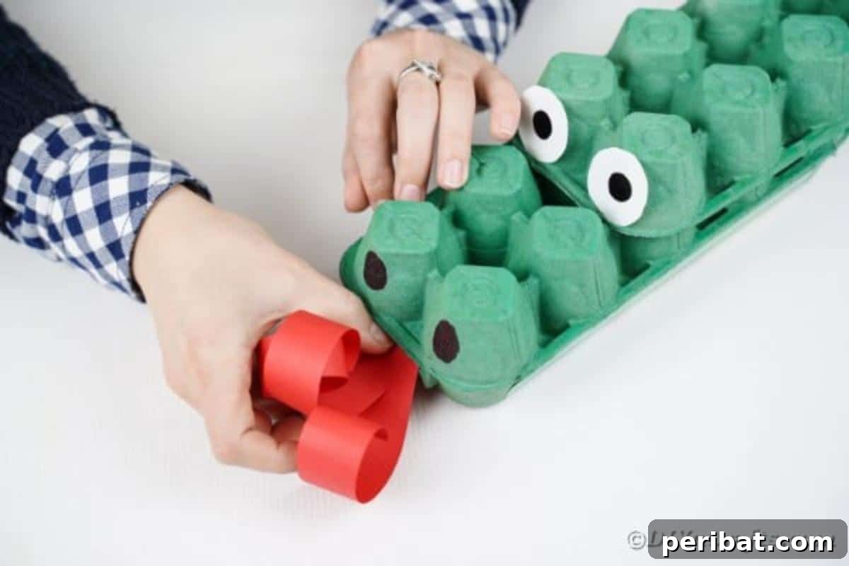 A hand gluing the curled red paper tongue onto the front of the green egg carton dragon's snout.