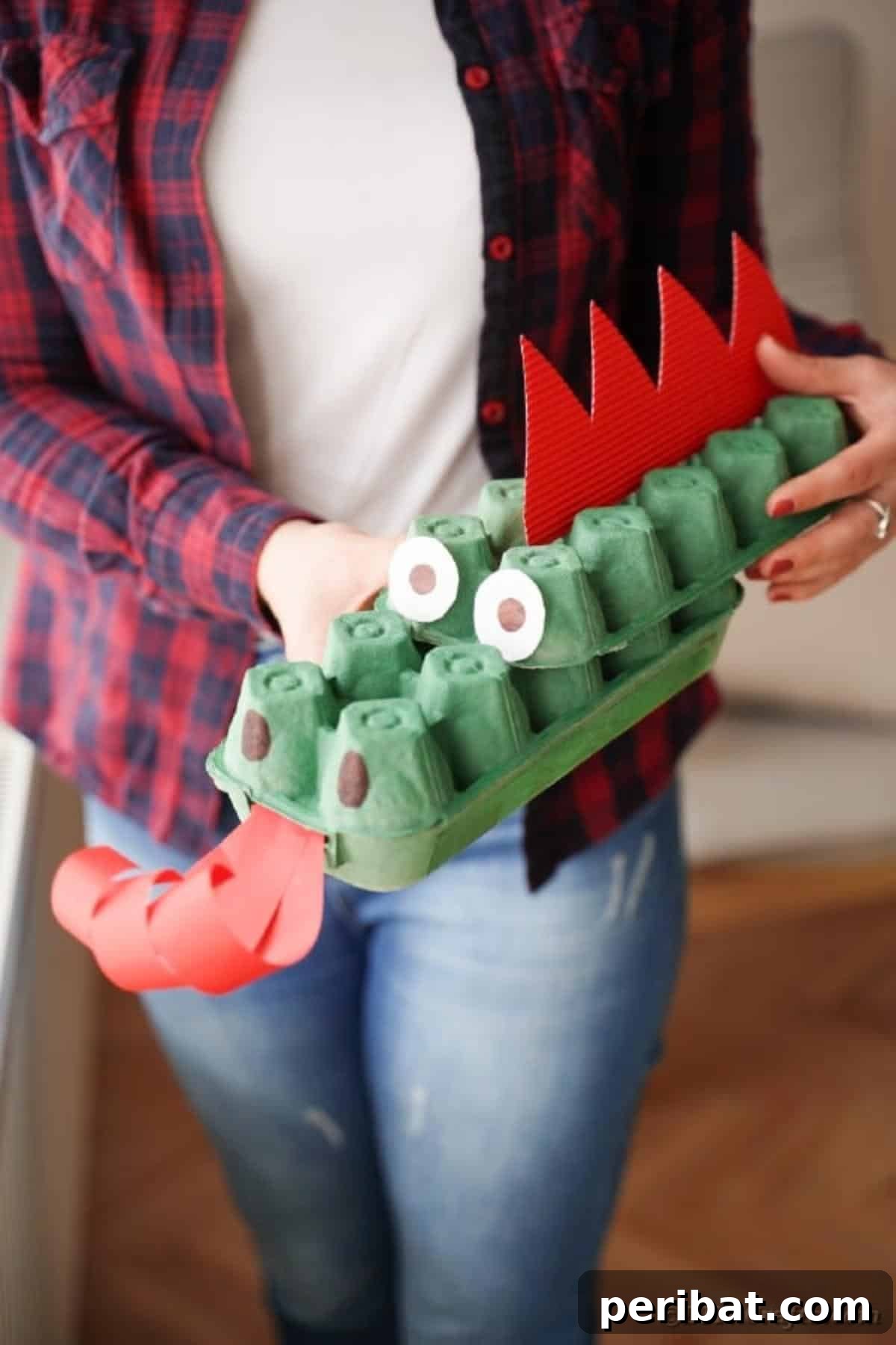 A woman holding a finished green egg carton dragon with red spikes and curled red tongue.