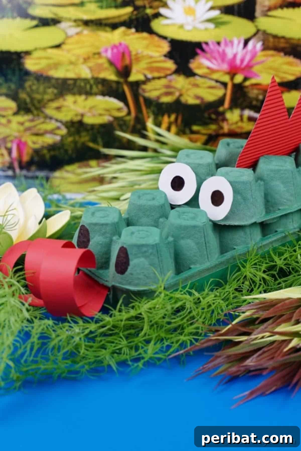 A green egg carton dragon with red details, set against a blurred natural background suggesting a pond or greenery.