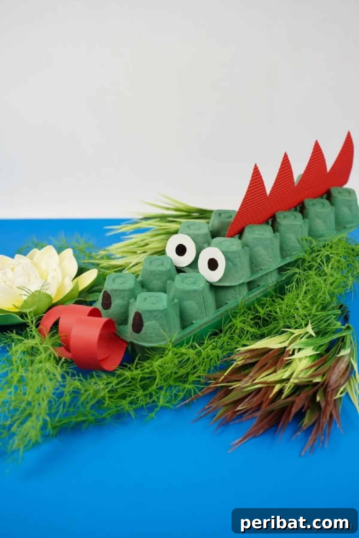 A detailed shot of a green egg carton dragon with expressive eyes and red spikes, on a textured blue surface.