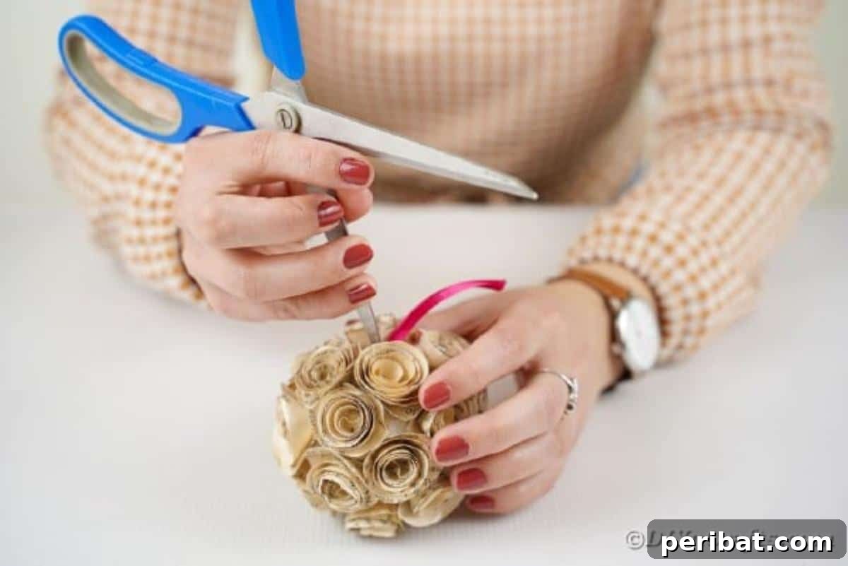 From Pages to Petals: DIY Book Rose Sphere 15 A hand sticking a ribbon hanger into the center of a paper rose ball, preparing it for display