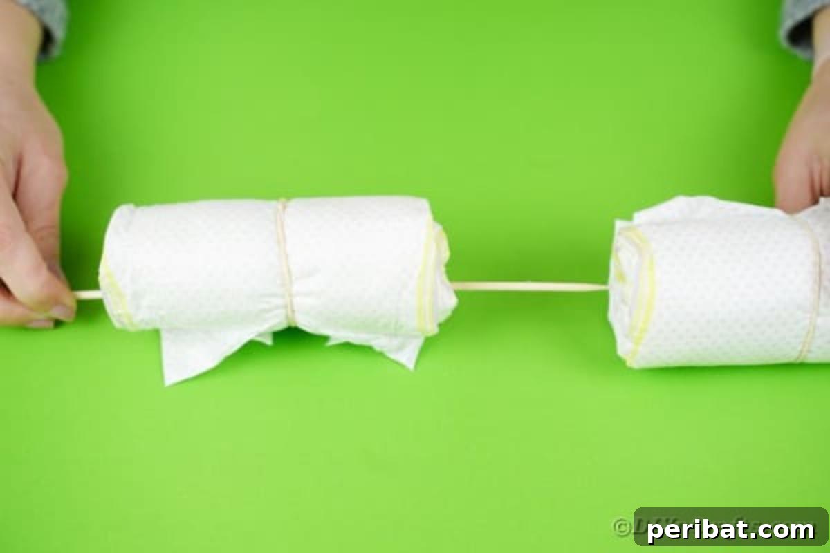 Two rolled diapers being carefully slid onto a wooden dowel, demonstrating how to create segments for the diaper crane's structural base.