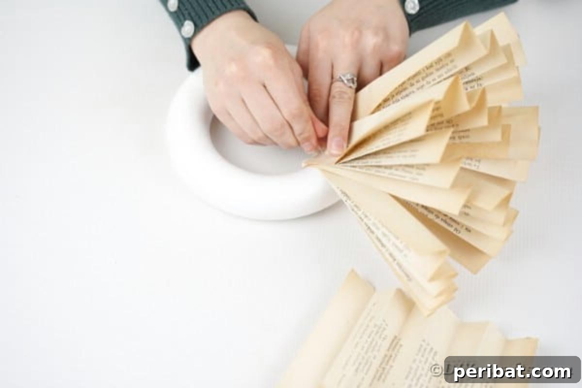 A person carefully attaching a folded paper fan from a book page to a Styrofoam wreath form with a push pin