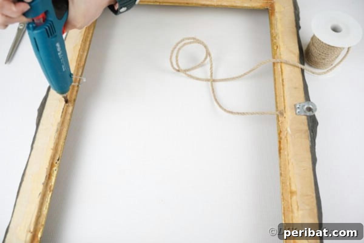 A person applying hot glue to the back of an ornate picture frame to attach a piece of twine